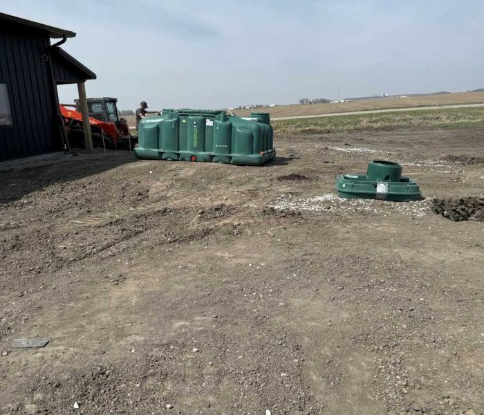 091525 DMX Gallery (2) A group of green plastic septic tanks sits on bare dirt near a dark building and orange utility vehicle on a rural construction site, with open fields and blue sky in the background.