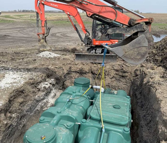 091525 DMX Gallery (3) An orange Kubota excavator lowers two large green plastic tanks into a trench at a construction site in a rural area, with dirt, fields, and blue sky in the background.