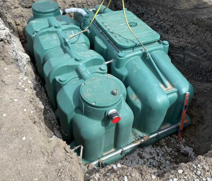 091525 DMX Gallery A large green septic tank is installed in a dug-out pit in the ground at a construction site, with pipes, wires, and surrounding dirt visible.