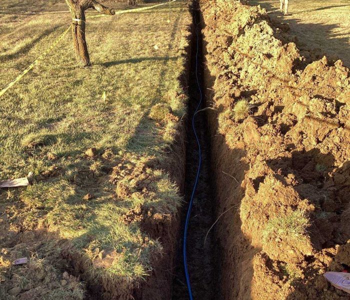2ca655a5-c76e-4a23-b0a3-265eef8cfc55 A deep, narrow trench runs through a grassy yard toward a house. A blue pipe lies at the bottom of the trench, and a person stands near the house in the background. Piles of dirt border both sides of the trench.