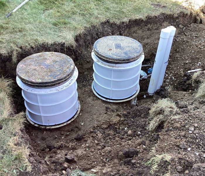 2e0a8950-fbfe-46ec-8df6-7c1bd16204de Two white plastic septic tank risers with metal lids are partially buried in the ground, surrounded by disturbed soil and grass; a white vent pipe stands nearby, and a shovel lies on the lawn.