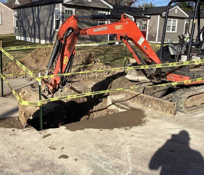 3fbd6024-934d-46e3-8c79-8bb3e1d473bf A small orange excavator digs a large hole in a residential street, surrounded by yellow caution tape. Nearby houses and trees are visible in the background, and a person's shadow appears in the foreground.