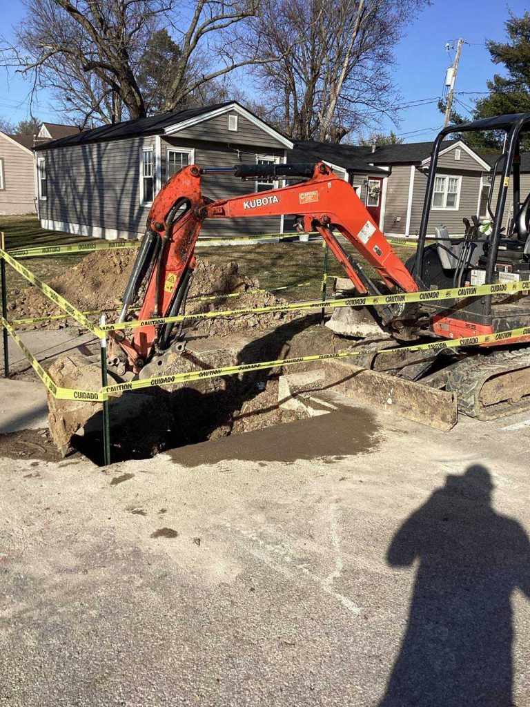 A small orange excavator digs a large hole in a residential street, surrounded by yellow caution tape. Nearby houses and trees are visible in the background, and a person's shadow appears in the foreground.