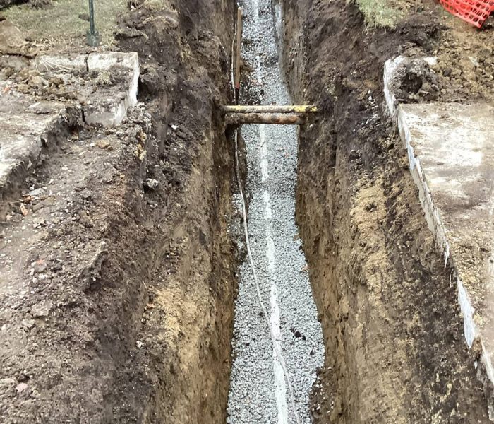 4940e9e9-28d3-4065-91b4-a7cc9295baeb A deep, narrow trench filled with gravel runs through soil, bordered by orange safety fencing and patches of grass, with a utility pipe and a wooden brace crossing the trench.