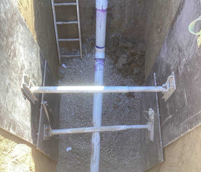 57b738df-6848-40ed-986a-ea4367c0c929 A trench with gravel at the bottom contains a white PVC pipe running horizontally, supported by two metal braces. Large black boards line the trench walls, and a ladder leans against one side.