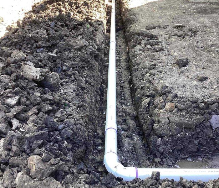 99e4c647-e233-4d00-94bb-9d12a0871200 A white PVC pipe is laid in a trench dug through soil, running parallel to the ground and turning at a right angle. The surrounding area is covered with loose, dark earth and appears to be part of a plumbing installation.