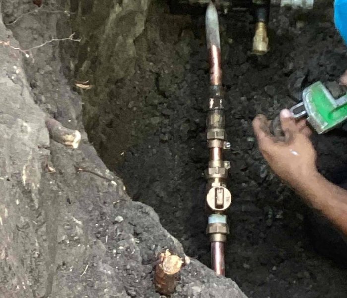 f3f073eb-976d-4714-a2d8-3f78077239d0 A person works in a dirt hole, using a green tool to repair or install copper and metal pipes connected by valves and fittings underground.