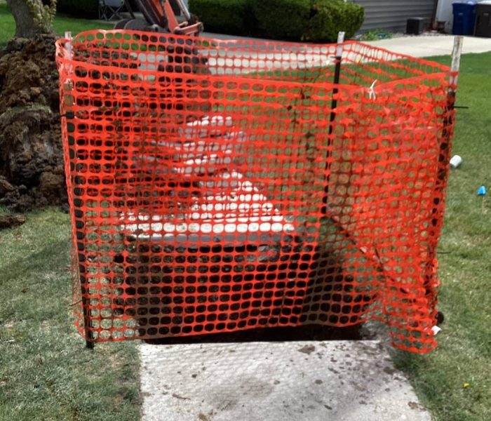 f9c52537-9535-4602-91fb-1d0fb8483fe4 A sidewalk section is blocked off with bright orange safety fencing for construction work. An excavator is digging nearby, and the fenced area contains dirt and debris in front of a house.