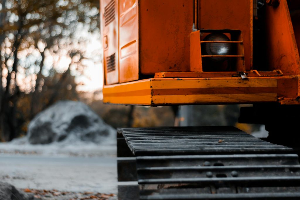 Close-up of the side and metal tracks of an orange construction excavator, with a blurred background of trees and a gray rock outdoors.